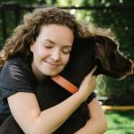 Smiling female with closed eyes embracing pointing dog in collar while sitting on lawn in summer